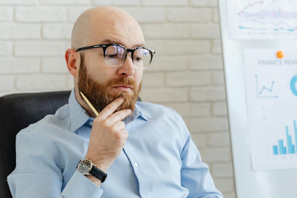 Bearded man in blue shirt contemplating financial data on whiteboard indoors.