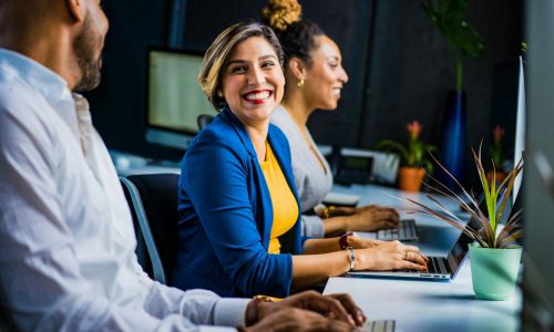 Three diverse professionals working and smiling at office desks, fostering teamwork and collaboration.