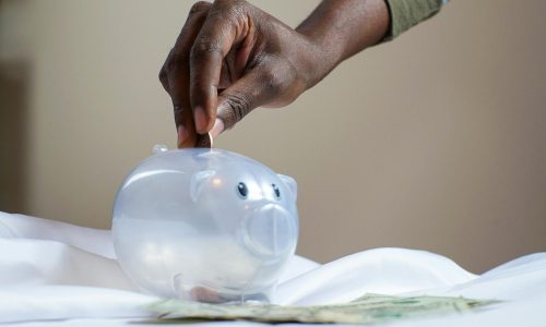 Close-up of a person's hand placing coins into a transparent piggy bank to save money.