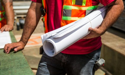 Construction workers in safety vests holding blueprints at a site during the day.