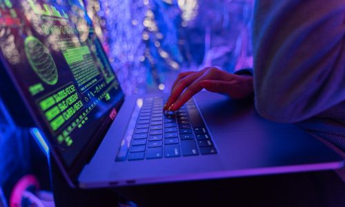 Close-up of hands typing on a laptop displaying cybersecurity graphics, illuminated by purple light.