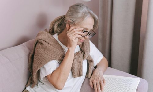 Elderly woman with glasses reading on a couch for leisure at home.