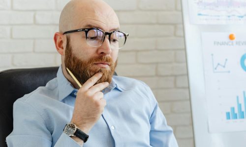 Bearded man in blue shirt contemplating financial data on whiteboard indoors.